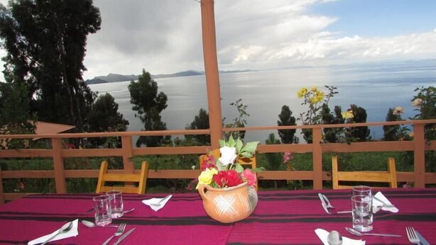Una mesa con mantel rojo, un florero y vista a un lago con una valla de madera al fondo.