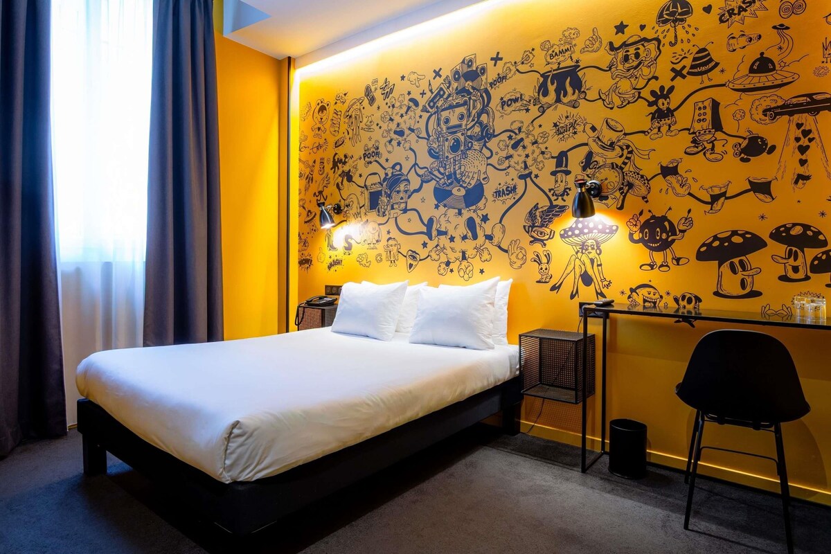 A hotel room with a bed, a desk, and a chair. The room has a yellow wall with black doodle artwork.