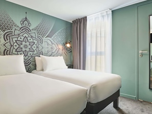 Una camera d'albergo con due letti, una parete decorata, una finestra con tende e una porta.
