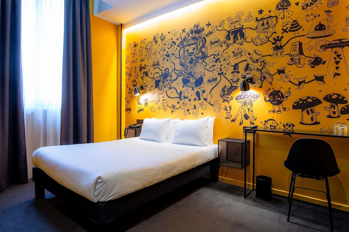 Habitación de hotel con una cama, un escritorio y una silla. La pared es amarilla y tiene dibujos artísticos estilo doodle.