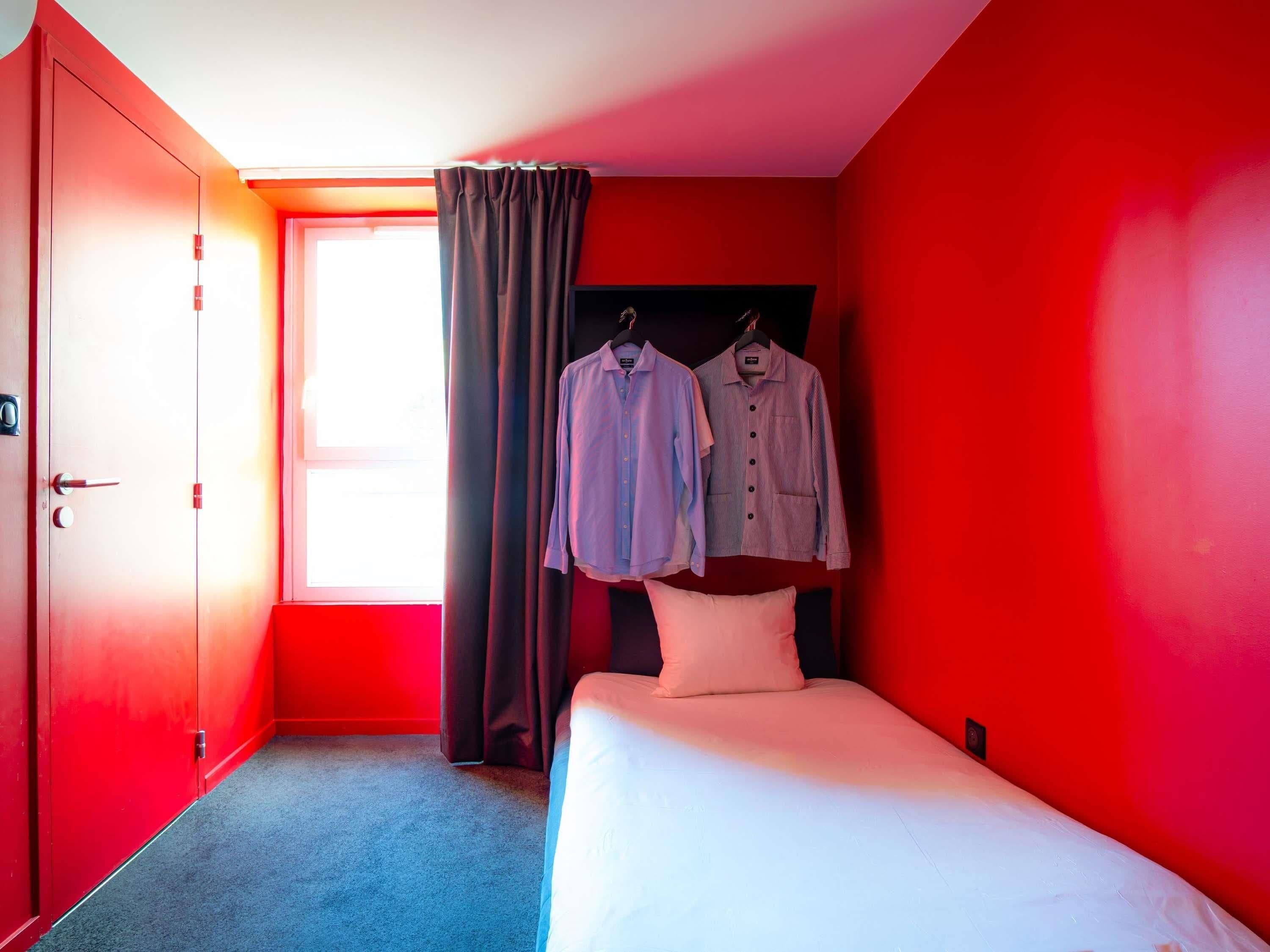 Une chambre d’hôtel avec un lit, deux chemises suspendues, une fenêtre avec des rideaux et un mur rouge.
