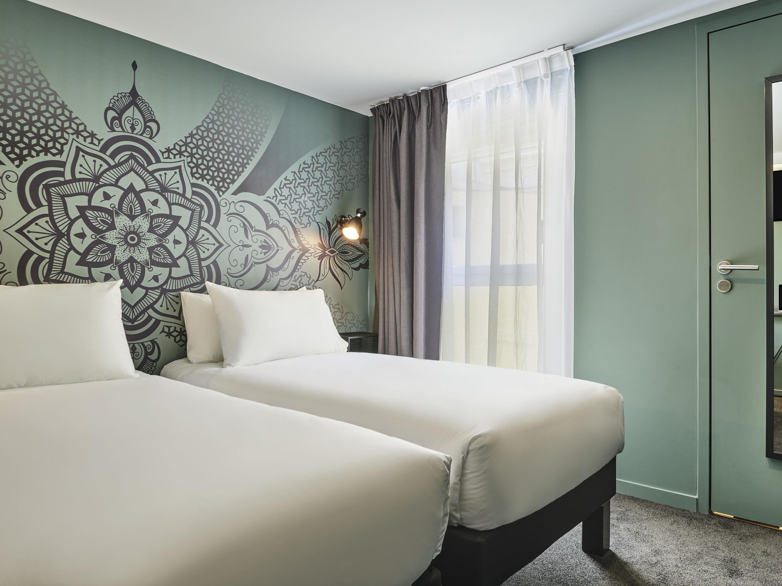 Ein Hotelzimmer mit zwei Betten, einer Wand mit Muster, einem Fenster mit Vorhängen und einer Tür.