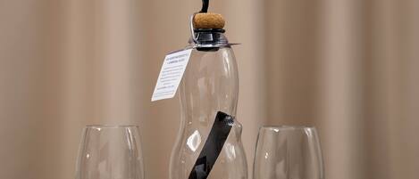 A glass bottle with a cork stopper and a black label, placed between two empty glasses on a wooden table.