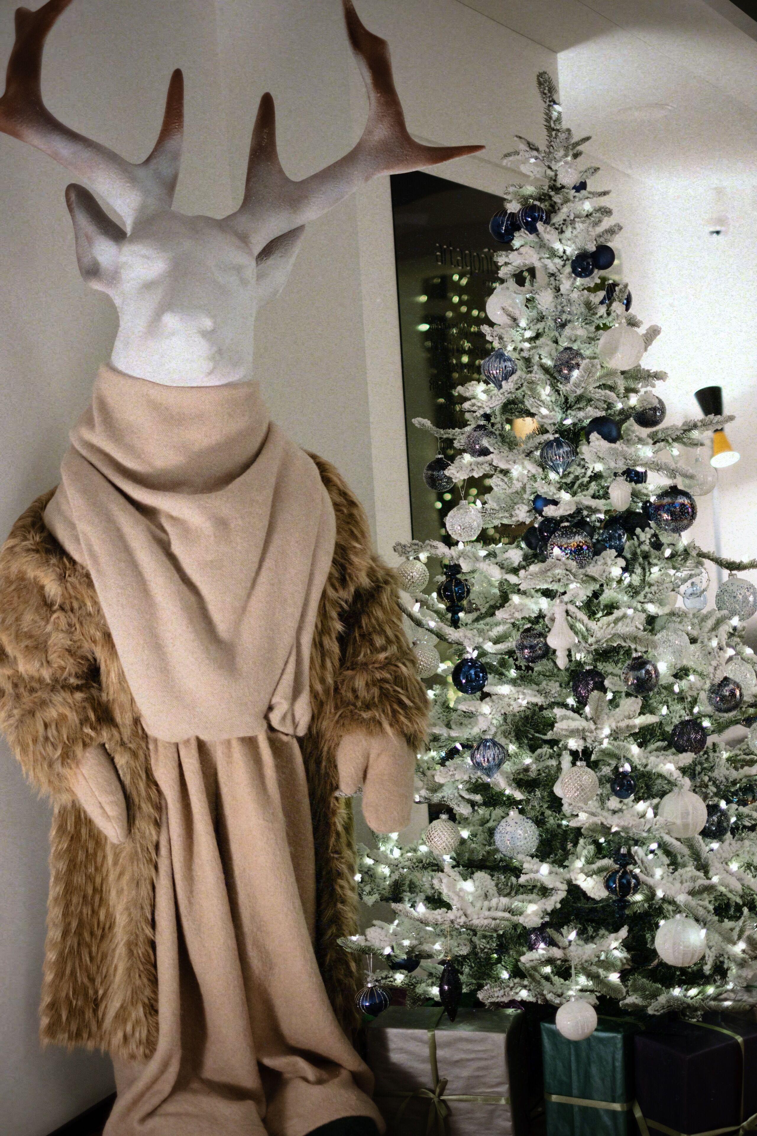 A mannequin with a deer head and a fur coat, a white Christmas tree with ornaments, and wrapped gifts.