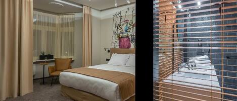 Ein Hotelzimmer mit einem großen Bett, einem Schreibtisch mit Stuhl und Blick durch ein Fenster mit Jalousien.