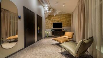A hotel room with a brick wall, a flat-screen TV, a sofa, a chair, a mirror, and a carpeted floor.