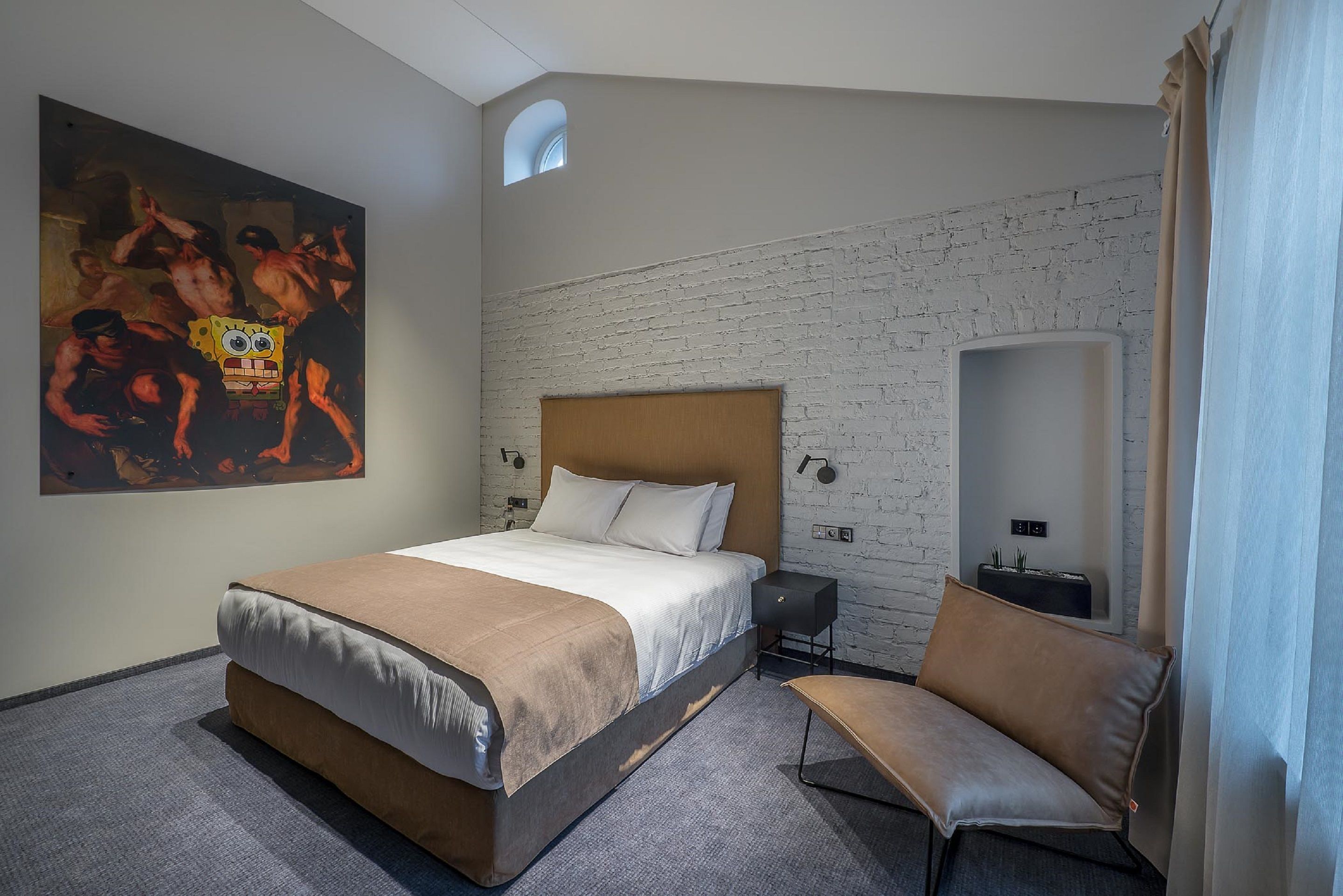 Une chambre d’hôtel avec un grand lit, une chaise, un tableau, un mur en briques et une petite fenêtre.