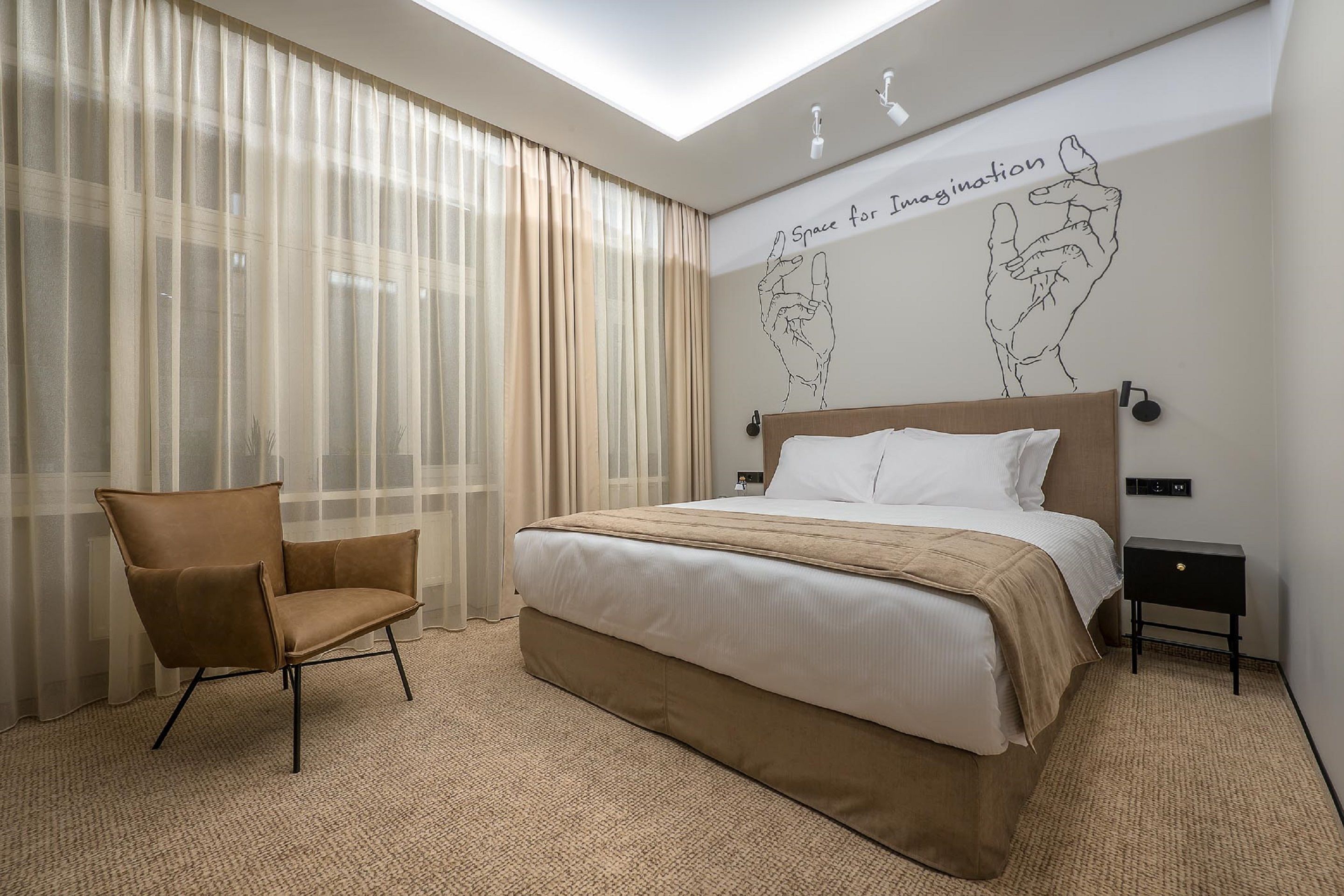 Een moderne hotelkamer met een groot bed, een stoel, een nachtkastje en een muur met een handgetekende illustratie en tekst.