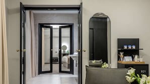 Habitación de hotel con cama, zona de estar con una silla y una mesa, espejo, estante con objetos y puerta que da a otra habitación.