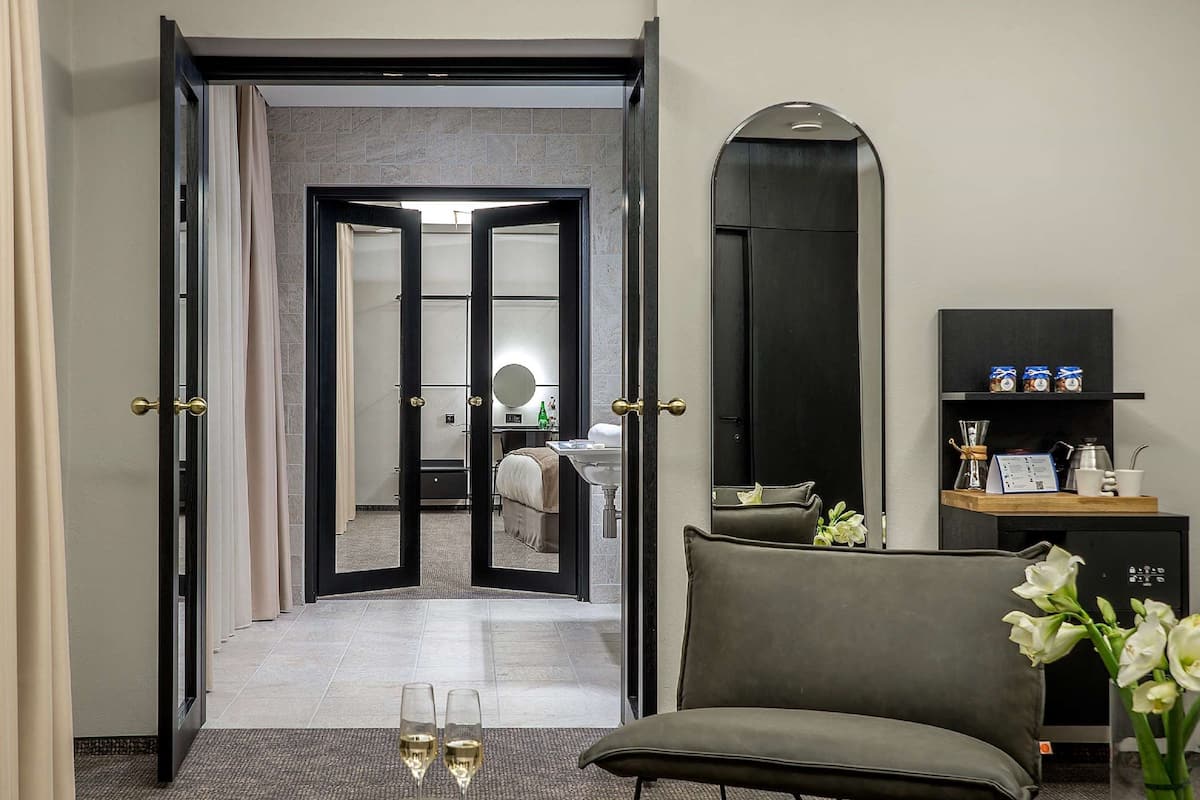 A hotel room with a bed, a sitting area with a chair and a table, a mirror, a shelf with items, and a door leading to another room.