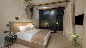 Una habitación de hotel moderna con una cama grande, mesita de noche y un espejo. Hay una pequeña mesa con una lámpara y un jarrón de flores. La habitación tiene una pared decorativa oscura y un techo de madera.