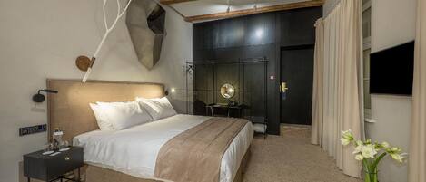A modern hotel room with a large bed, bedside table, and a mirror. There is a small table with a lamp and a vase of flowers. The room has a dark accent wall and a wooden ceiling.