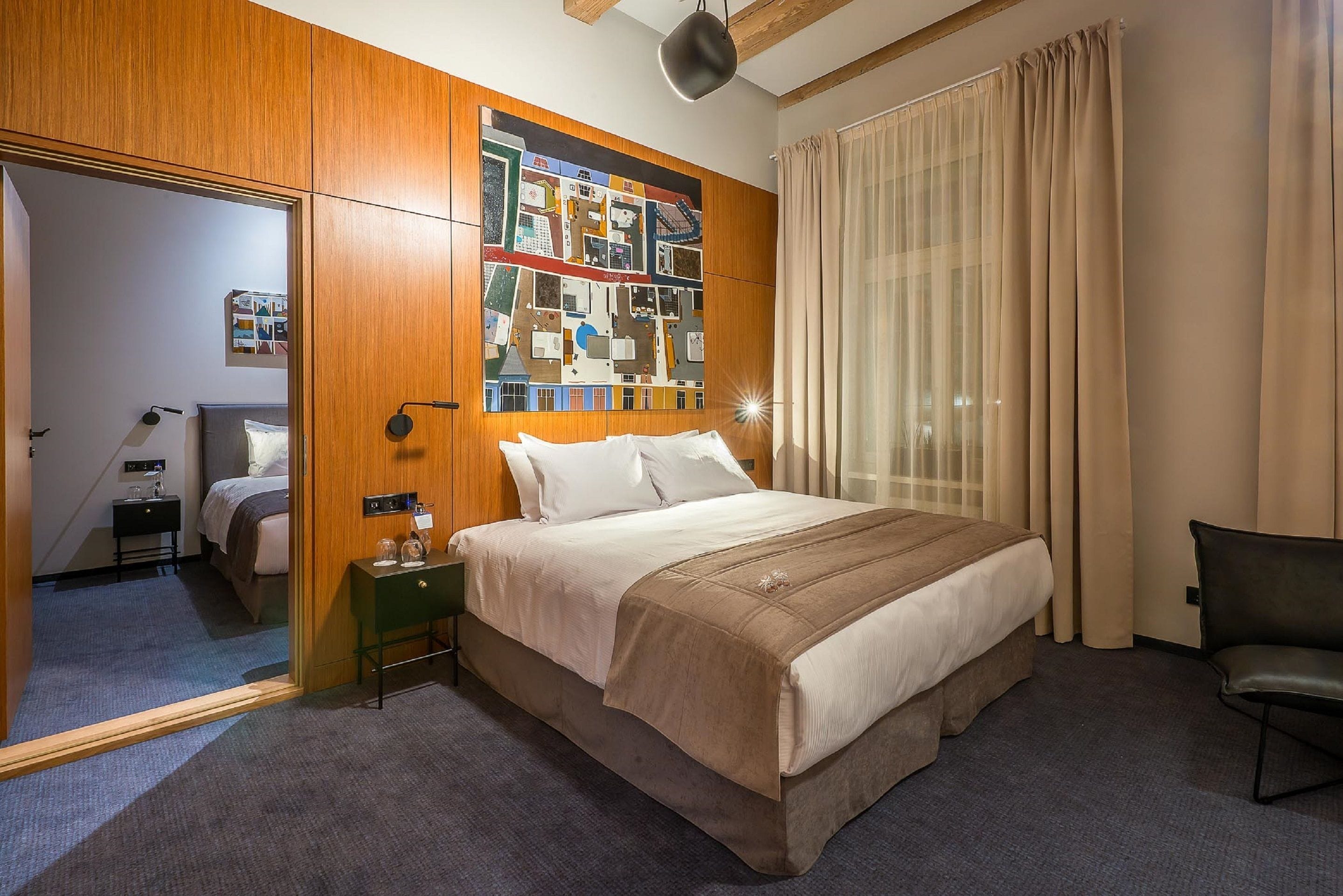 A hotel room with a large bed, a wooden wardrobe, a nightstand, a chair, and a wall art piece.
