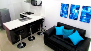 A compact kitchen with a microwave, sink, and gas stove. There is a black sofa with blue cushions and a wall-mounted air conditioner.