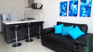 A modern kitchen with a white countertop, a stove, and a microwave. There are three black bar stools and a black leather sofa with blue cushions.