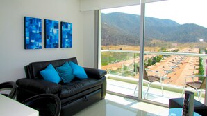 A modern living room with a black leather sofa, two blue abstract paintings, and a large balcony with a view of a residential area and mountains.