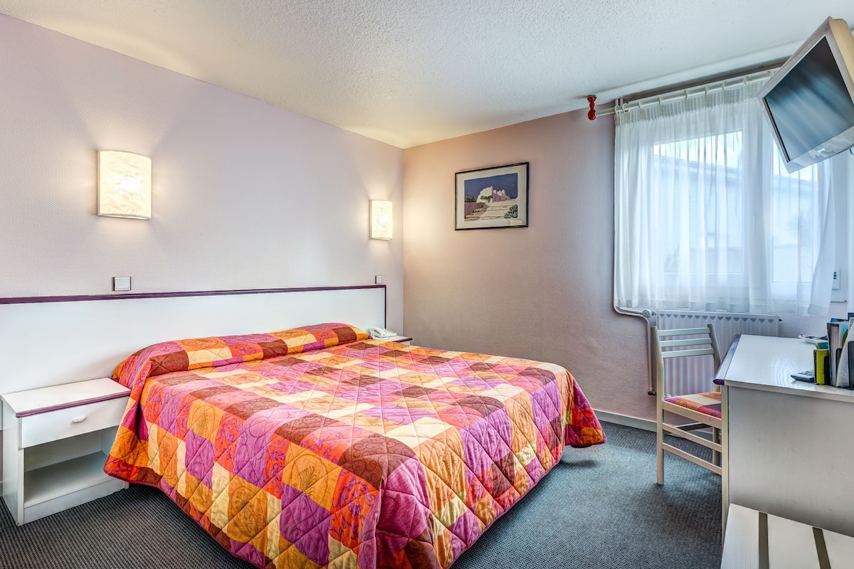 Une chambre d’hôtel avec un lit, un bureau, une chaise, une télévision et une fenêtre avec des rideaux.
