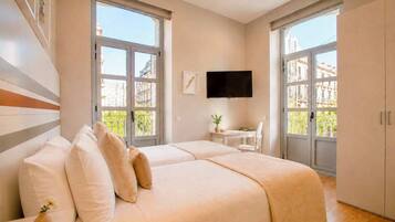 A hotel room with two beds, a TV, a small table with a chair, and large windows.