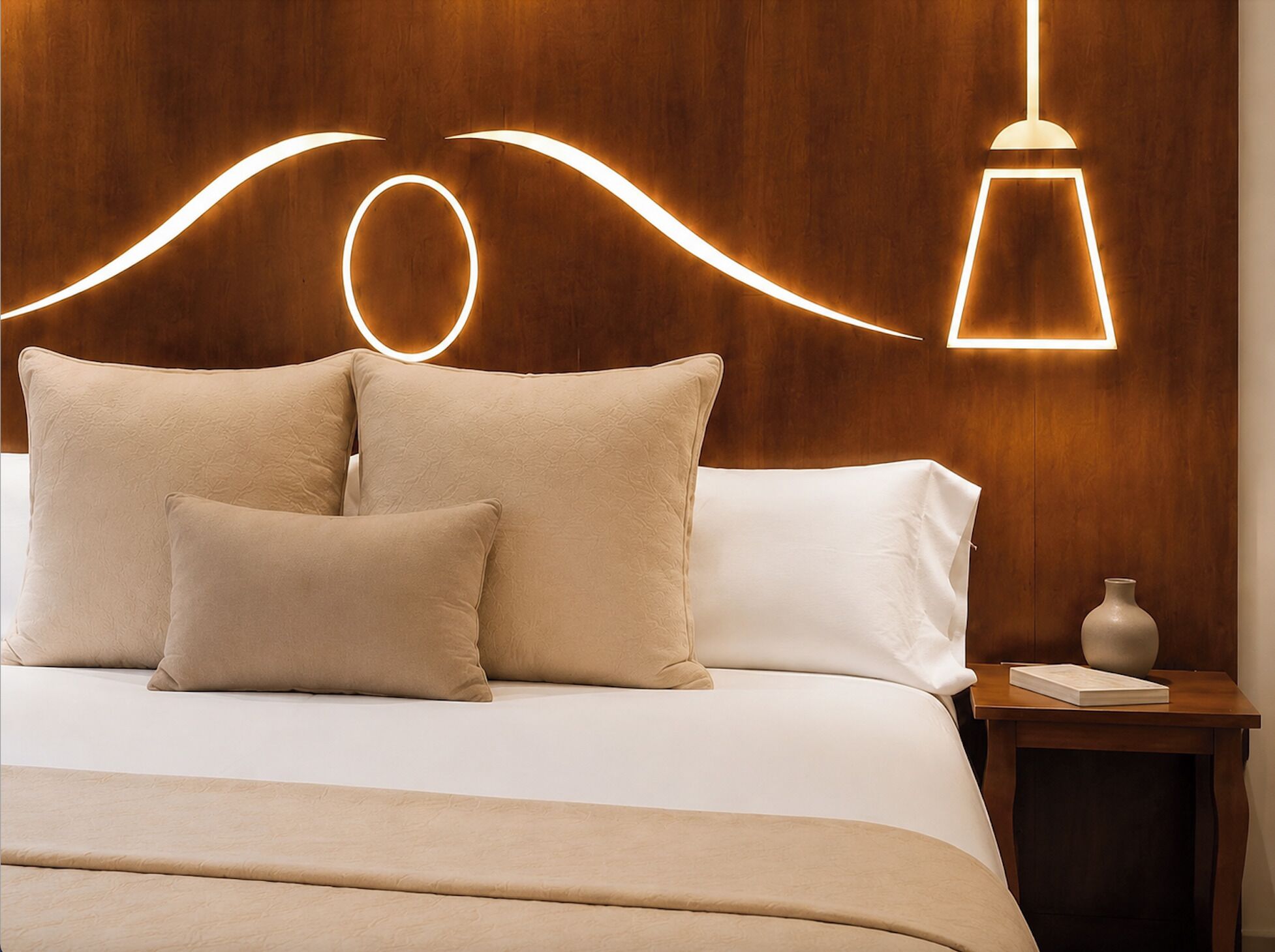 A neatly made bed with a wooden headboard, two pillows, a bedside table with a vase and book, and a modern pendant light.