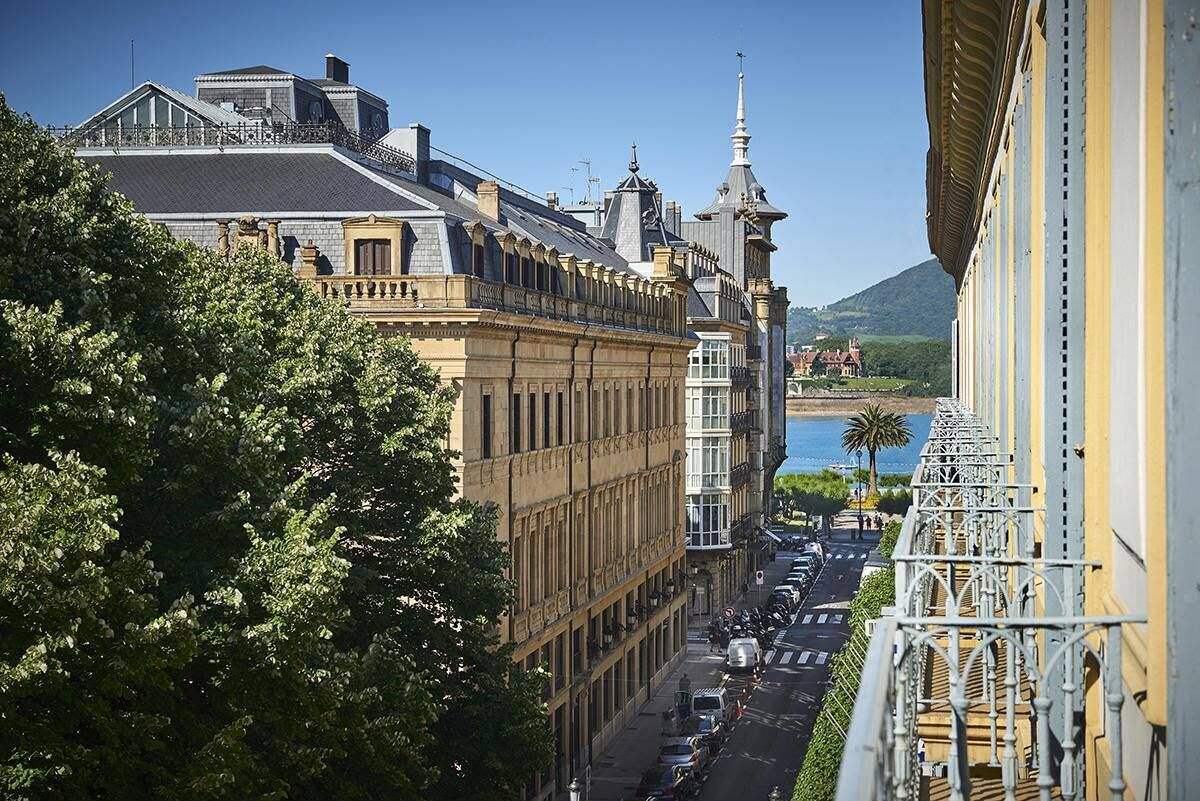 Un edificio histórico con arquitectura clásica, un balcón con barandillas ornamentadas y vistas a una calle con coches aparcados y una montaña distante.