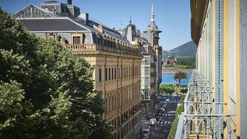 Un edificio histórico con arquitectura clásica, un balcón con barandillas ornamentadas y vistas a una calle con coches aparcados y una montaña distante.