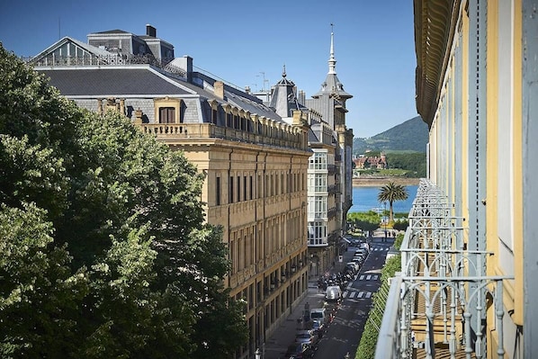 A historic building with classical architecture, a balcony with ornate railings, and a view of a street with parked cars and a distant mountain.