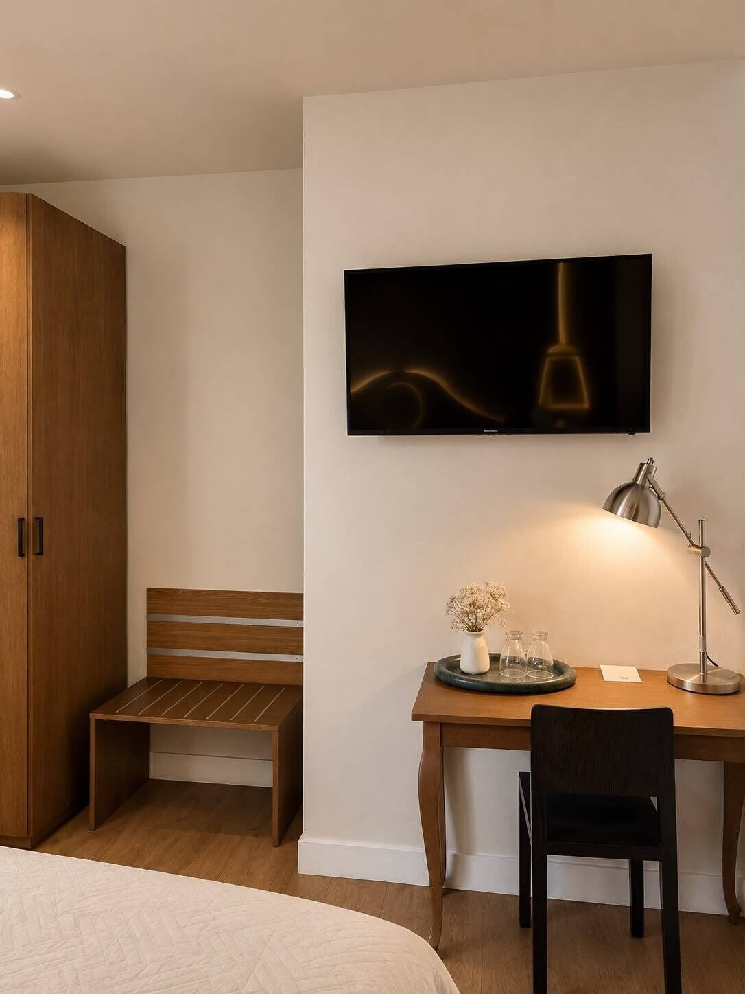 A bedroom with a bed, a desk with a chair, a wooden wardrobe, and a wall-mounted TV.