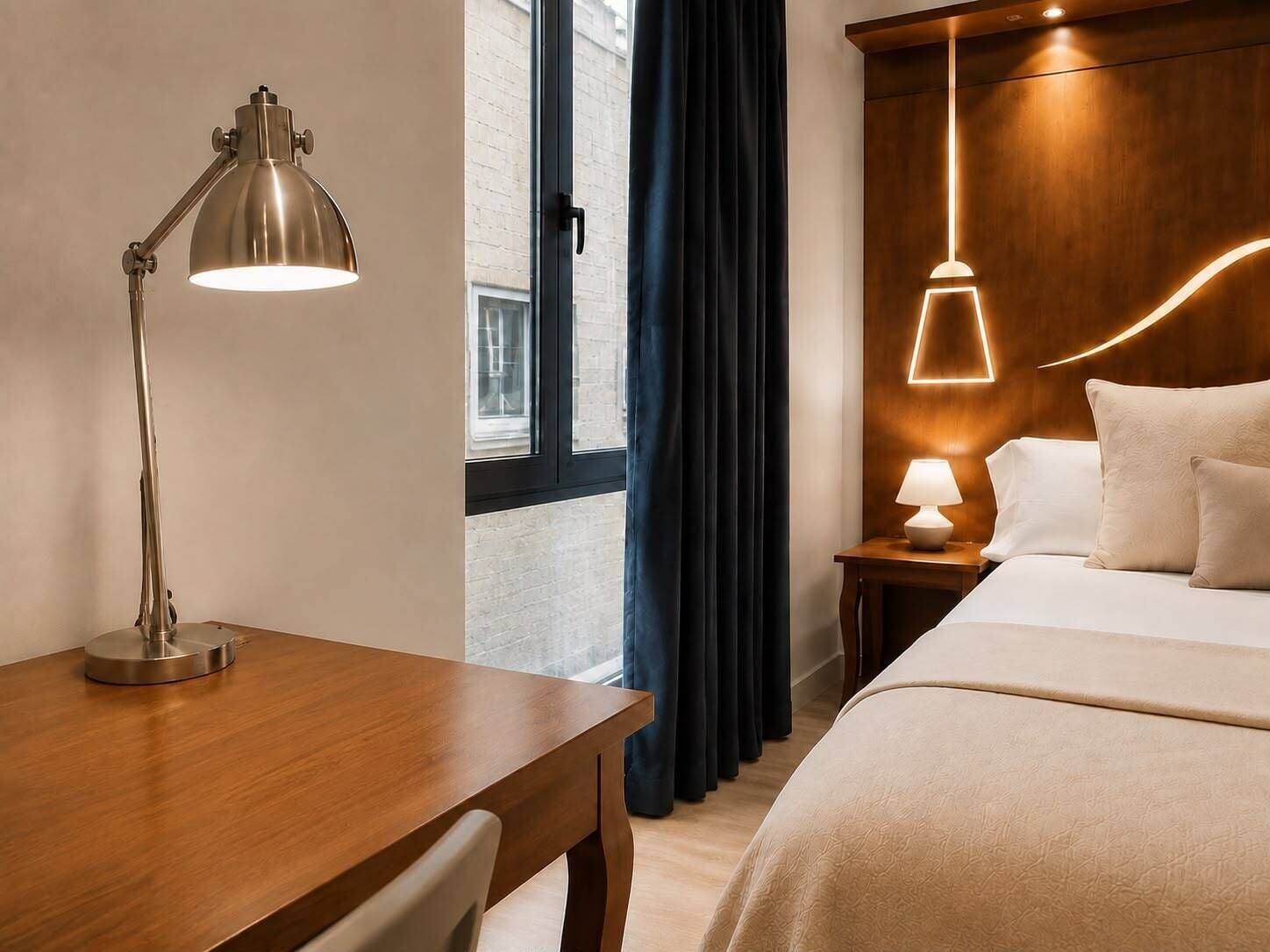 Habitación de hotel con un escritorio de madera, una lámpara, una cama con ropa de cama blanca, una mesita de noche y un aplique en la pared.