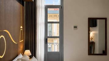 Habitación de hotel con una cama grande, una mesita de noche con lámpara, un espejo y vista a la calle desde una puerta.