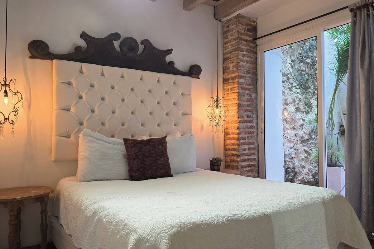 A bedroom with a tufted headboard, a bedside table, and a view of a brick wall and greenery outside.