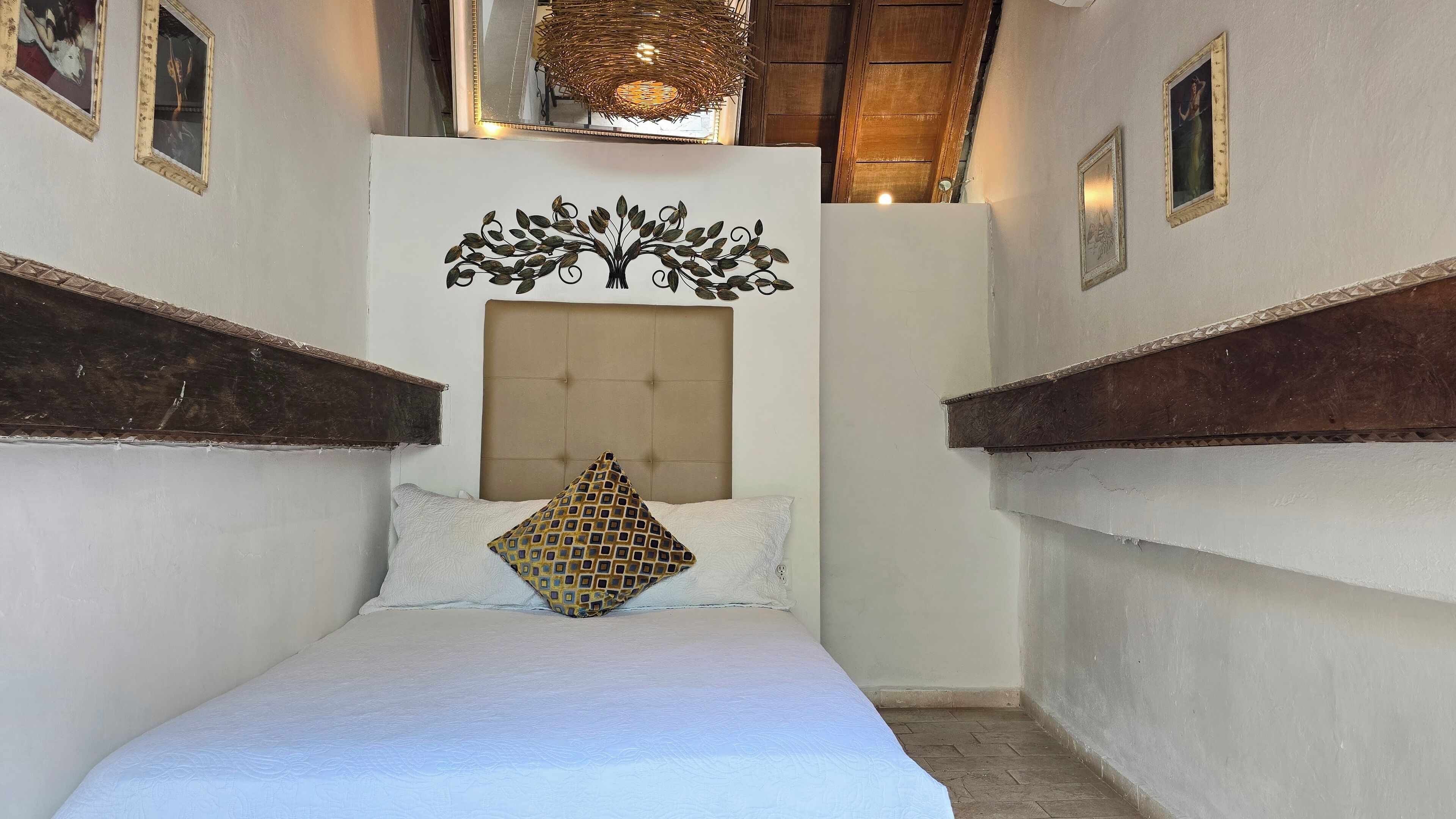 A narrow room with a bed, a decorative wall piece above the bed, and a wooden headboard.