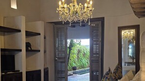 Un dormitorio con una cama grande, una araña de cristal, vigas de madera y vistas a un jardín a través de contraventanas abiertas.