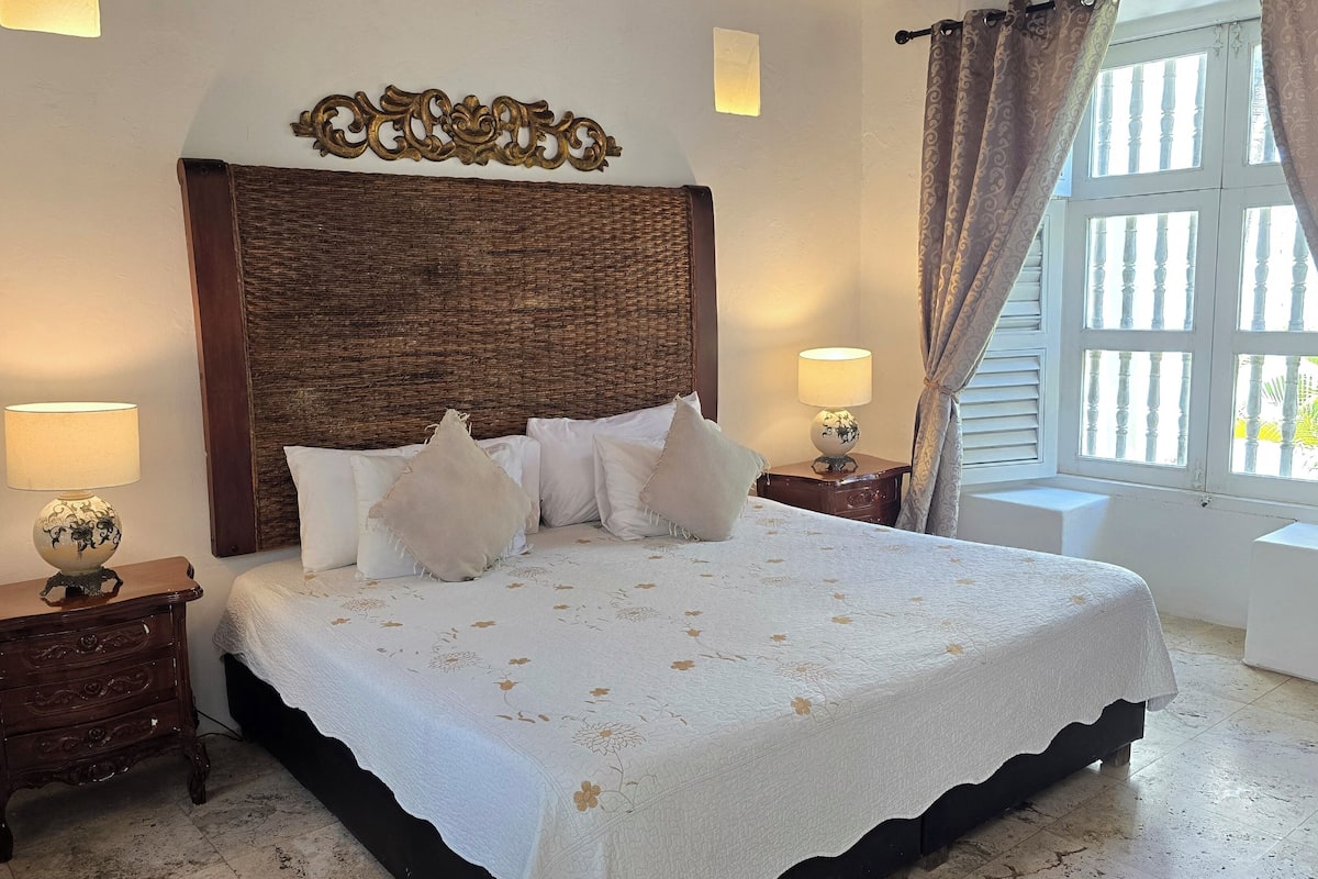 A bedroom with a large bed, two bedside tables, a decorative headboard, and a window with curtains.