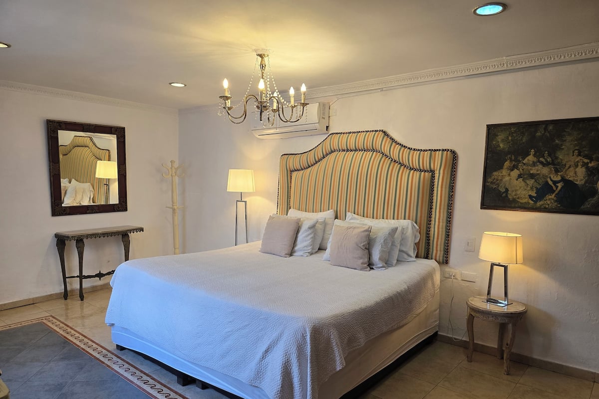 A spacious bedroom with a large bed, a chandelier, a painting, and a mirror.
