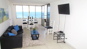 Een woonkamer met een zwarte bank, een salontafel en een televisie aan de muur. Op de achtergrond staat een eettafel met stoelen, en er is uitzicht op de oceaan door grote ramen.