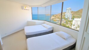 Een kamer met twee bedden, een groot raam met uitzicht op zee en gebouwen, en een airconditioning op de muur.