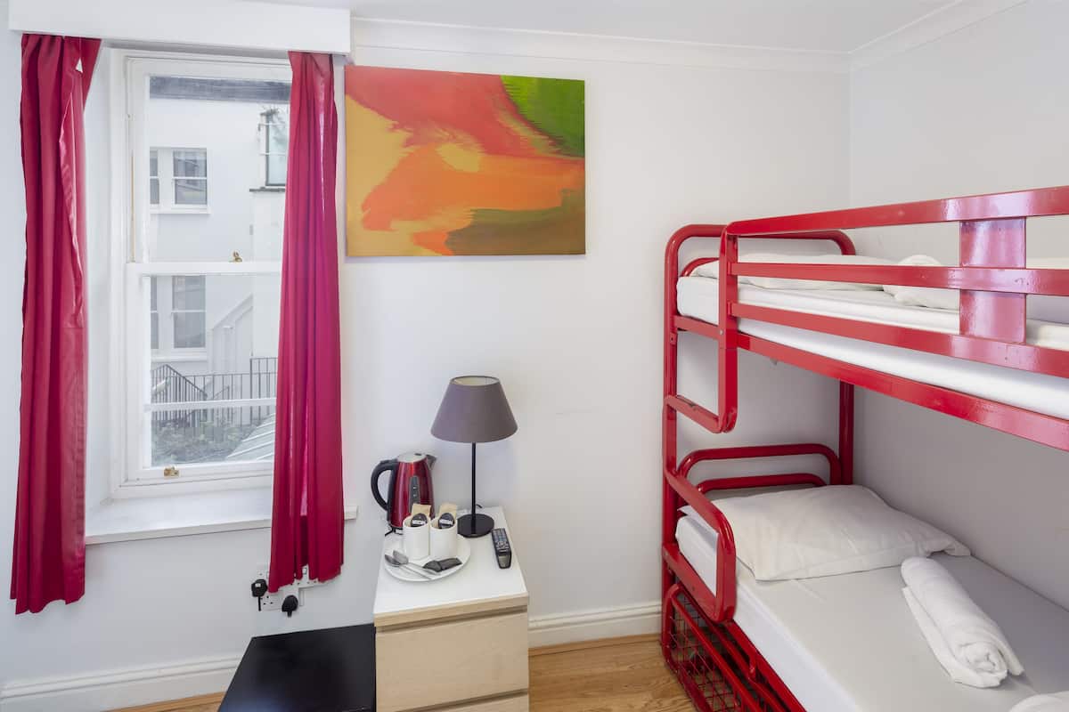 A bunk bed room with a window, a painting, a bedside table with a lamp, and a kettle.
