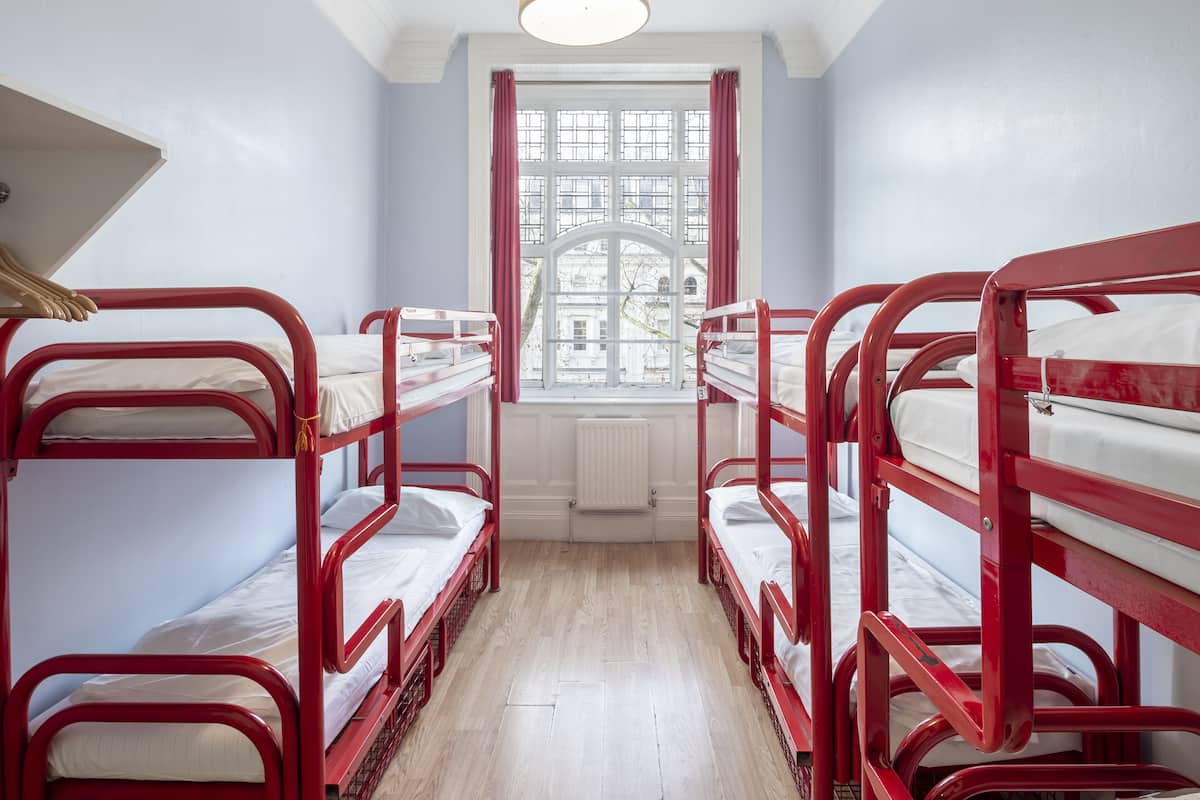 A dormitory room with four bunk beds, a radiator, and a window with curtains.