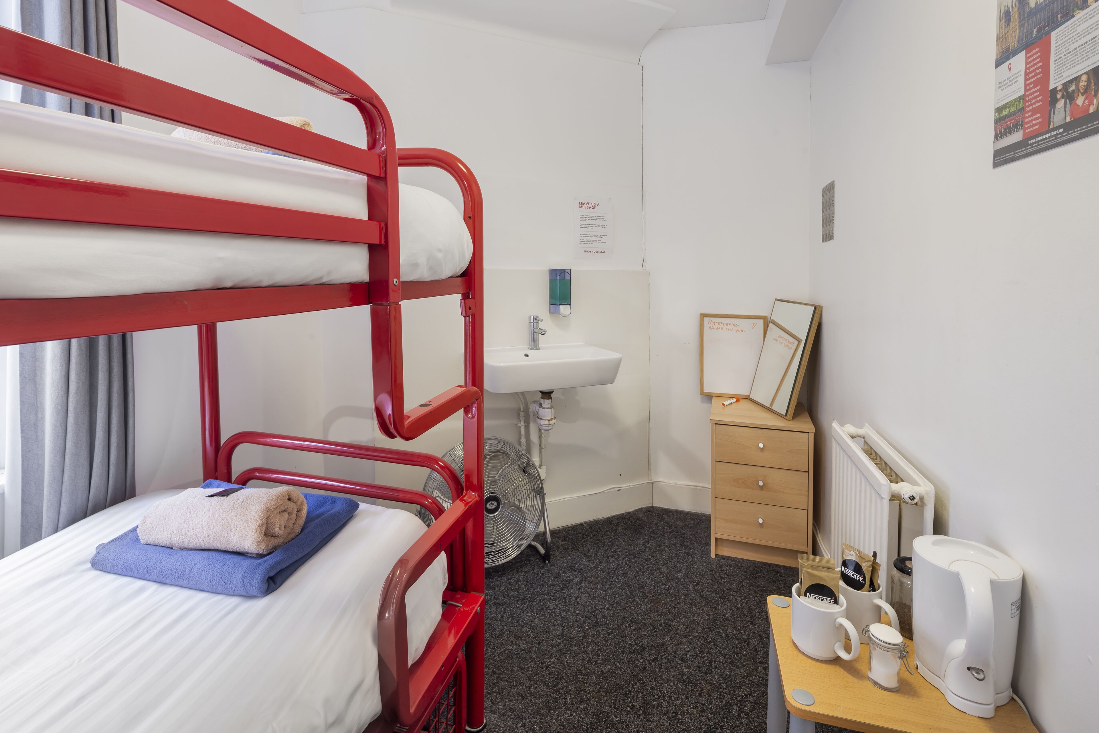 A bunk bed with a blue towel and a white pillow, a white sink, a wooden dresser, a framed picture, a coffee maker, and a fan.