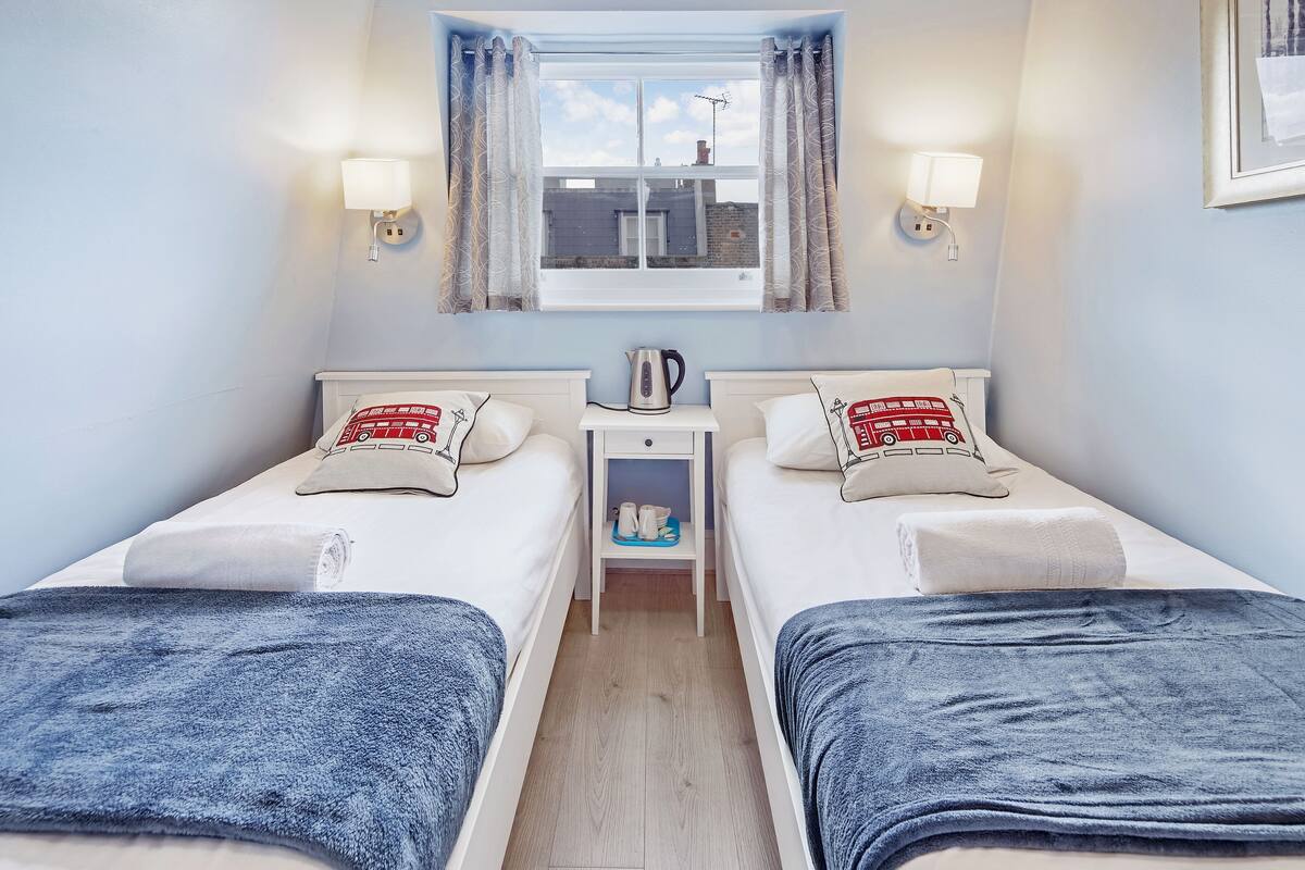Ein Doppelzimmer mit zwei Betten, einem Nachttisch, einem Wasserkocher und einem Fenster mit Vorhängen.