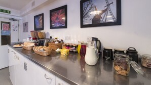 A breakfast bar with bread, pastries, and beverages. There are framed pictures on the wall and a sign indicating the direction to the exit. - Astor Kensington Hostel (London)