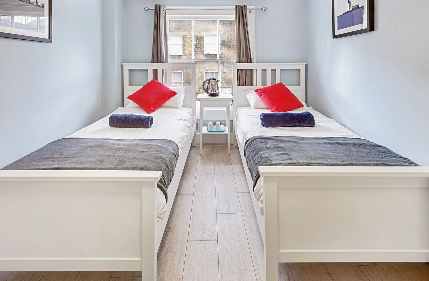 A twin bedroom with two beds, each with a red pillow and a grey blanket. There is a nightstand between the beds with a lamp and a small table.