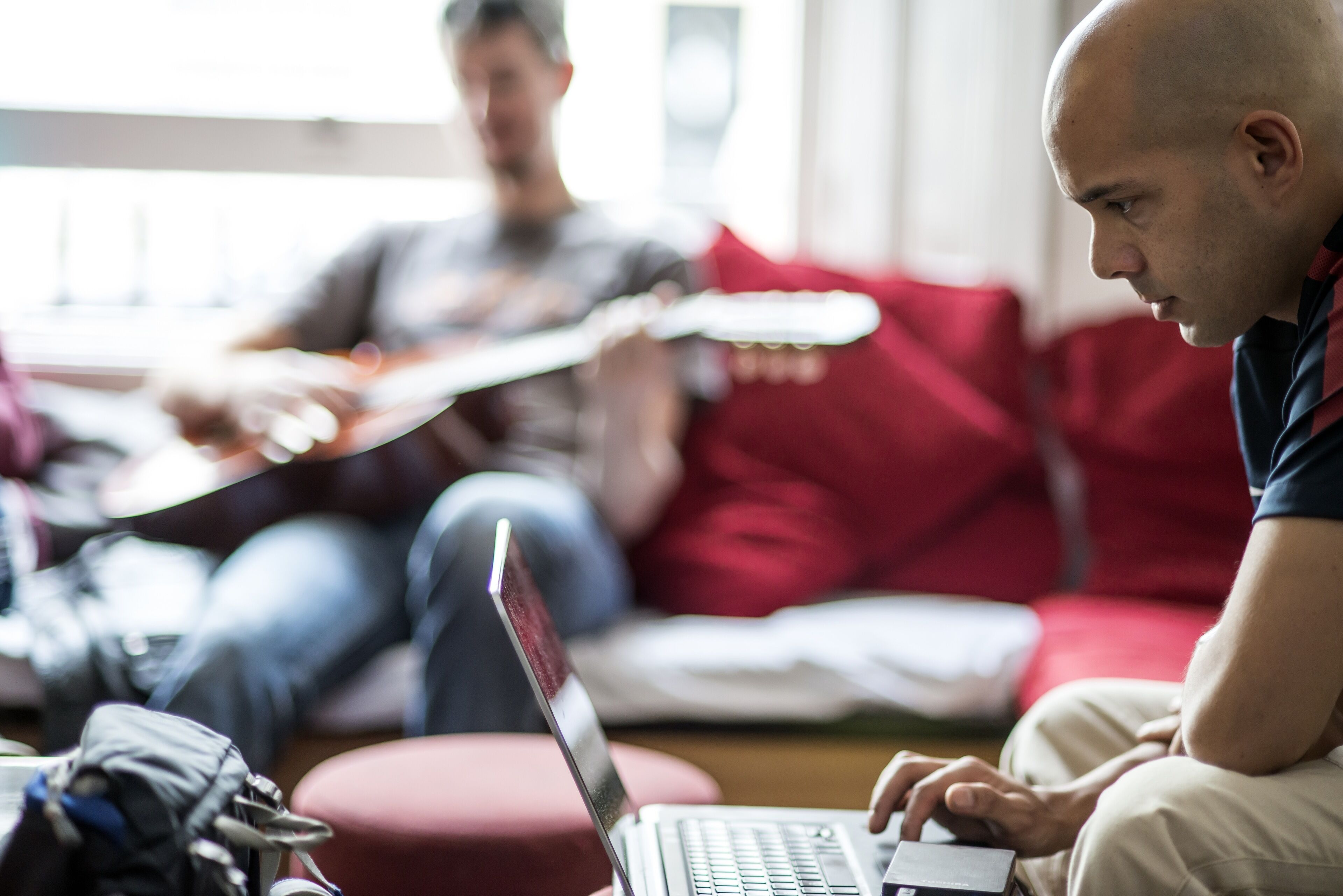 Eine Person benutzt einen Laptop auf einem roten Sofa in einem Raum, im Hintergrund steht eine Gitarre.