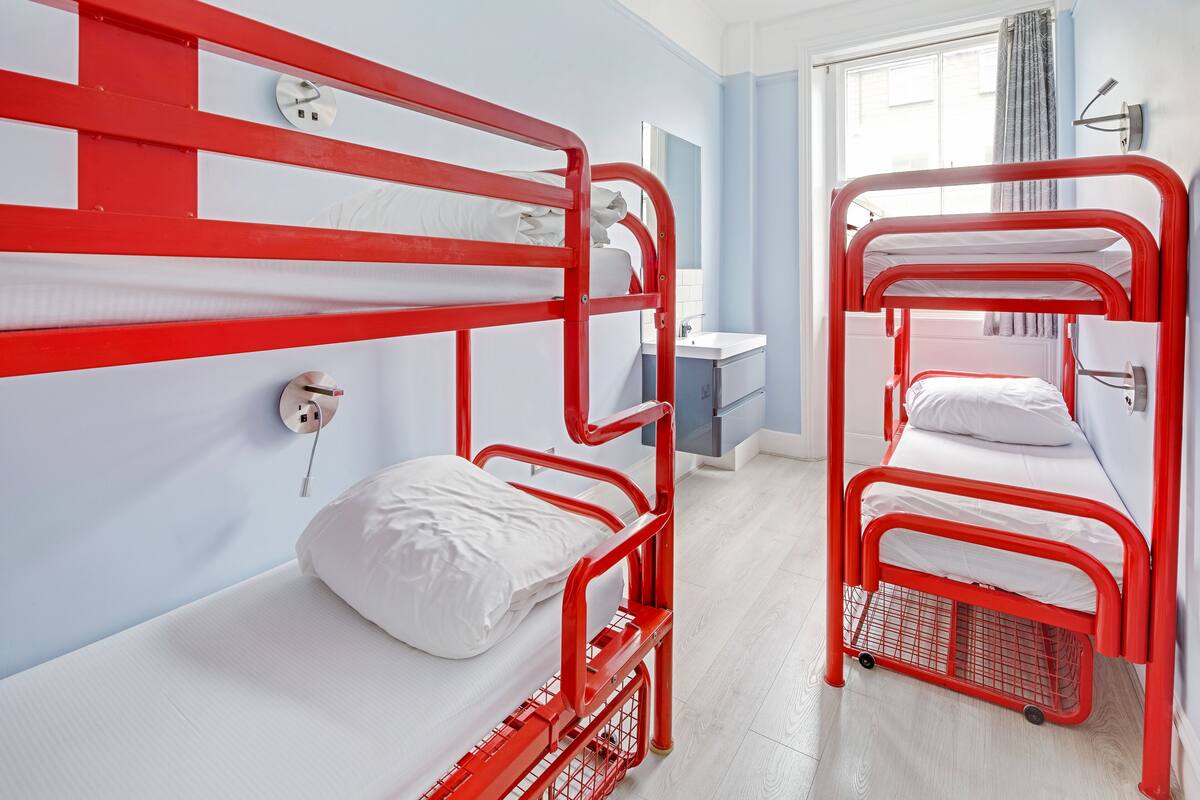 Ein Zimmer mit zwei roten Etagenbetten, einem weißen Nachttisch, einem Spiegel und einem Fenster mit Vorhängen.