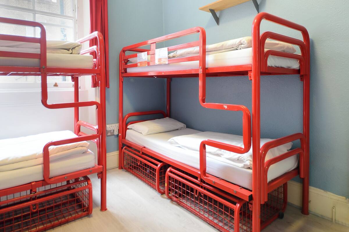 Ein Zimmer mit Etagenbetten, einem Fenster mit Vorhängen und einem Regal über den Betten.