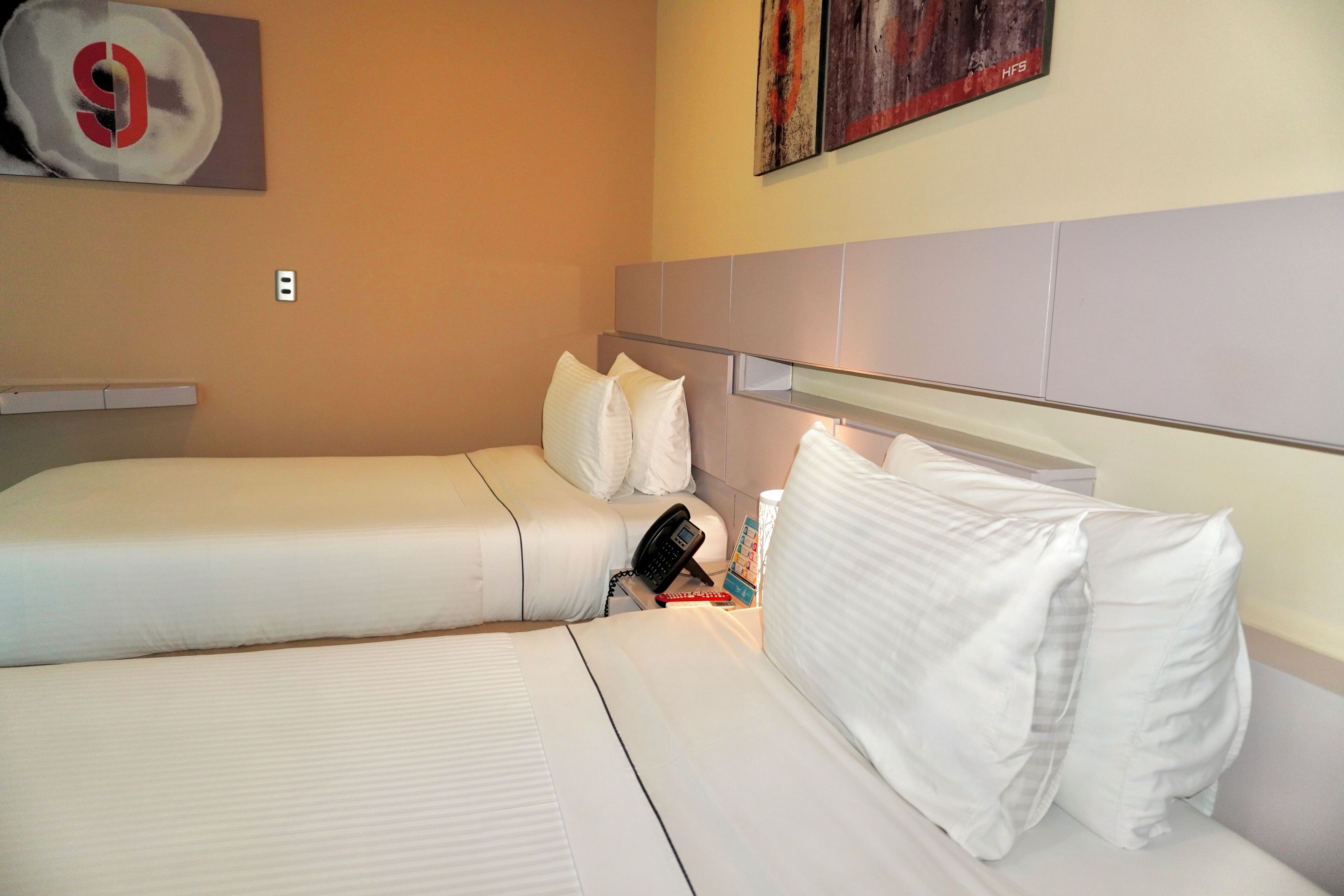Ein Hotelzimmer mit zwei Einzelbetten, einem Nachttisch mit Telefon, einer Lampe und einem an der Wand hängenden Kunstwerk.