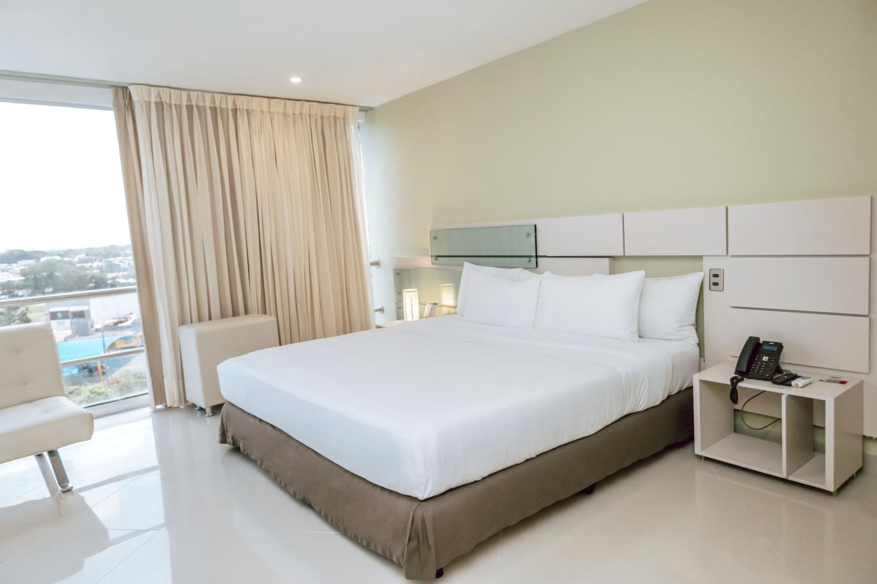 A modern hotel room with a large bed, a bedside table with a phone, and a view of the city through the window.