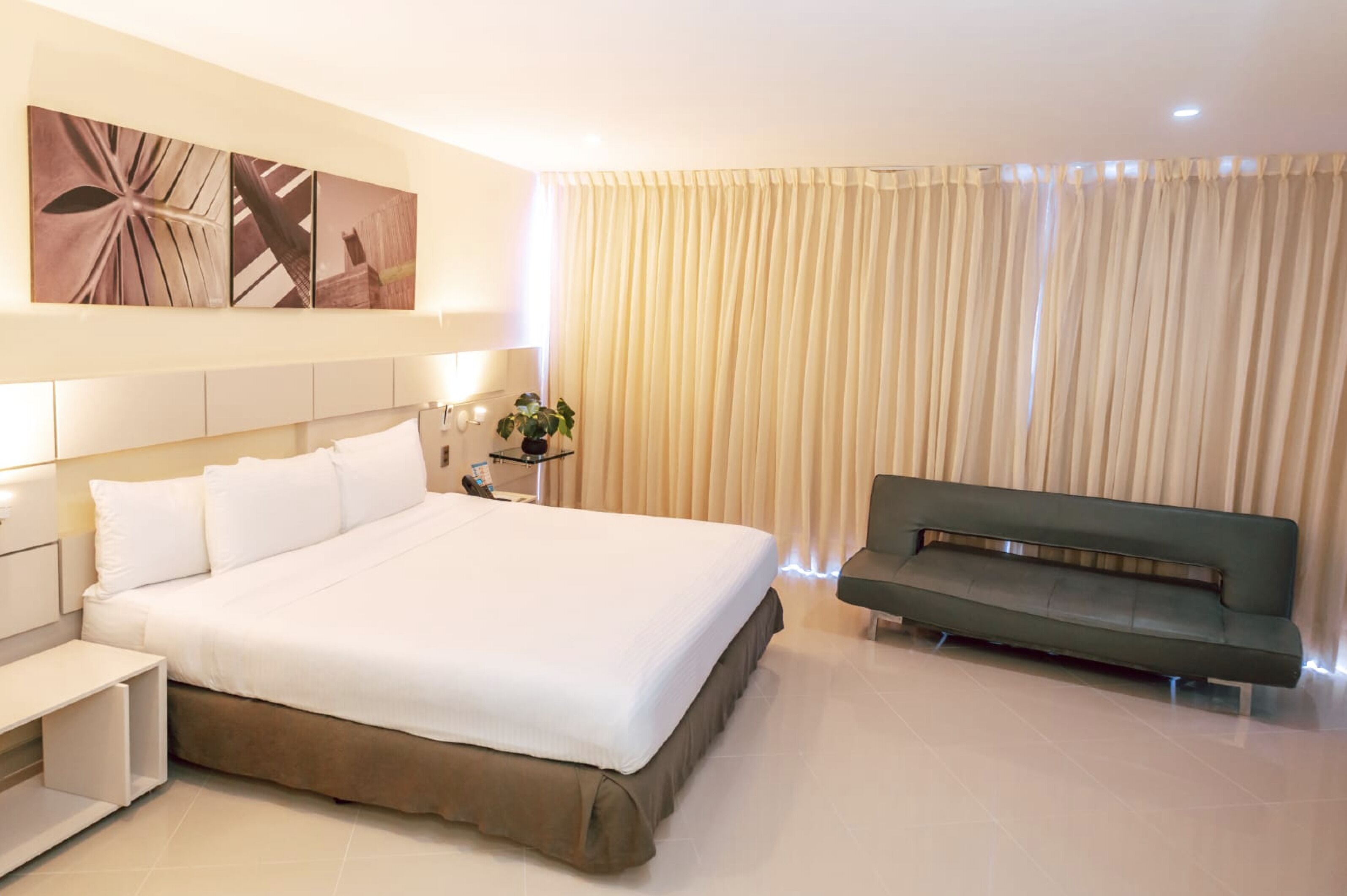 Una habitación de hotel con una cama grande, una mesita de noche, un sofá verde y un amplio ventanal con cortinas.