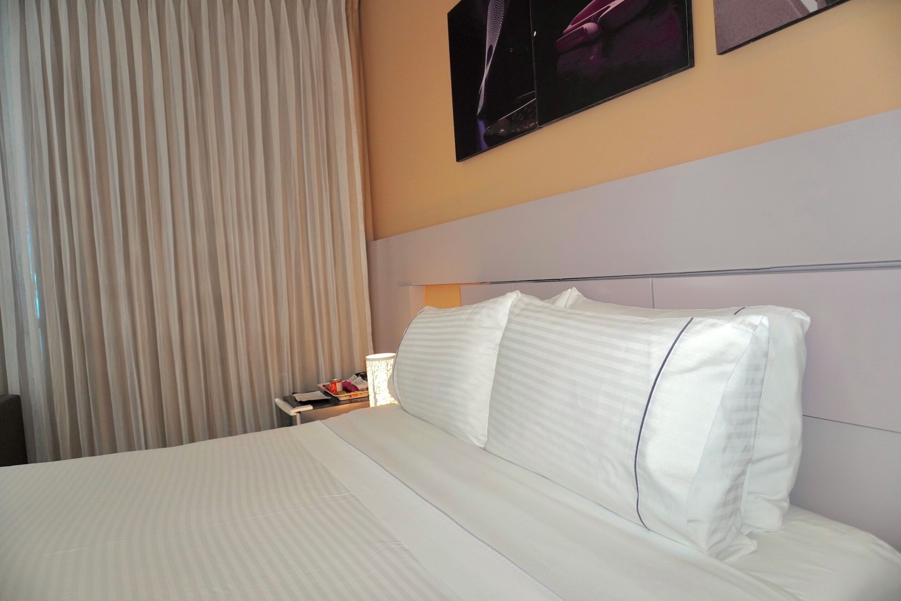 Ein Hotelzimmer mit Bett, Vorhängen, einem Nachttisch und einem an der Wand befestigten Fernseher.