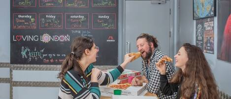 Three people sitting at a table with pizza boxes and a chalkboard wall displaying weekly events.